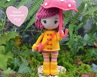 May include: A crocheted doll wearing a yellow raincoat, pink and white striped pants, and yellow rain boots. The doll is holding a pink and white mushroom umbrella. The doll is standing on a wooden base. The text "Patron uniquement PDF" is visible in the image.