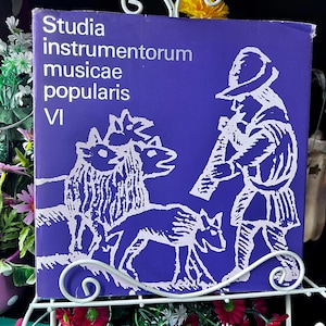 Könnte beinhalten: Ein quadratisches Buch mit violettem Einband und weißer Strichzeichnung einer Person, die Flöte spielt und neben einer Gruppe von Tieren steht. Der Text auf dem Einband lautet Studia instrumentorum musicae popularis VI in weißer Schrift links.