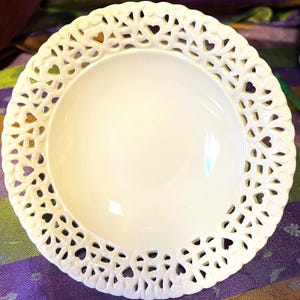 Puede incluir: Un plato de cerámica blanco con un diseño de encaje en forma de corazón alrededor del borde. El plato tiene una superficie lisa y brillante y una forma de cuenco poco profunda. Los cortes de corazón añaden un toque delicado al diseño.