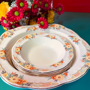 May include: A set of vintage white ceramic plates and bowls featuring a floral design in orange, yellow, and green. The plates have a scalloped edge and a brown trim. A bouquet of red flowers is in the background.