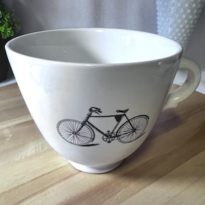 May include: A large, off-white ceramic mug with a black bicycle graphic. The mug has a wide, rounded bowl shape and a curved handle. The bicycle illustration is detailed, showing the frame, wheels, and other components.