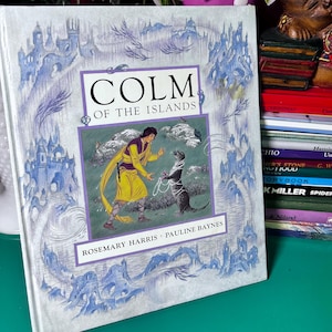 May include: A hardcover book titled "COLM of the Islands" with illustrations. The cover features a person in yellow clothing and a dark-colored animal, possibly an otter, holding a chain. The background has blue castle illustrations and the authors' names.
