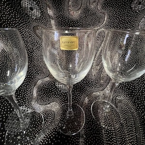 Op de afbeelding: Drie heldere Luminarc wijnglazen met lange stelen. De glazen hebben een klassiek ontwerp en een gouden label. De achtergrond is een zwarte stof met een zilver en wit patroon.