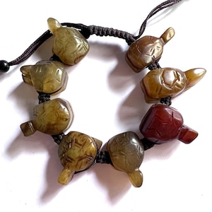 Puede incluir: Una pulsera con abalorios de tortuga tallados en tonos marrón, verde y rojo. Los abalorios están ensartados con un cordón marrón oscuro, creando un accesorio único. La pulsera se muestra sobre un fondo blanco.
