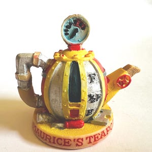 Könnte beinhalten: Eine skurrile Teekannenfigur im Steampunk-Stil. Auf dem gelben Sockel steht "MAURICE'S TEAPOT". Die Teekanne hat einen runden Körper mit roten, silbernen und gelben Akzenten, ein Messgerät oben und einen Ausguss mit rotem Ventil.