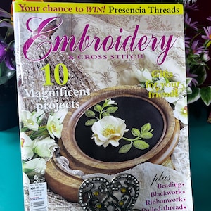 May include: A magazine titled "Embroidery Cross Stitch" with a floral design on the cover. The cover features a framed floral embroidery project. The magazine is propped up by a decorative heart-shaped stand. The text "Your chance to WIN!" is visible.