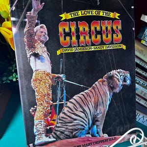 May include: Vintage book cover titled "The Love of the Circus" featuring a circus performer in a gold sequined outfit and a tiger. The cover has a black background with yellow and orange text. The book is propped up on a white metal stand.
