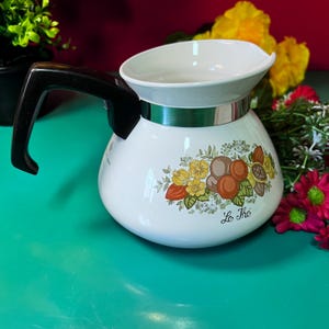 Puede incluir: Una taza de cerámica blanca con un asa negra y el texto "But First, Coffee" en negro, con una fuente de estilo manuscrito. La taza está sobre una superficie blanca.