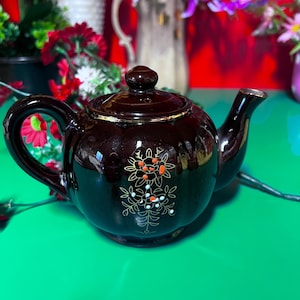 Op de afbeelding: Een donkerbruine keramische theepot met deksel en handvat. De theepot heeft een bloemmotief in oranje, wit en goud. De tuit en rand hebben gouden accenten. De theepot staat op een groene ondergrond.