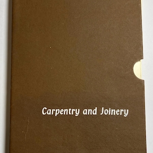 May include: A brown book with the title "Carpentry and Joinery" in white lettering. The book has a textured cover and a curved cutout on the right side, revealing a cream-coloured page.