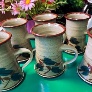 May include: A collection of six ceramic mugs with a speckled beige finish and a dark brown rim. Each mug has a handle and is decorated with blue and red leaf designs. The mugs are displayed on a green surface.