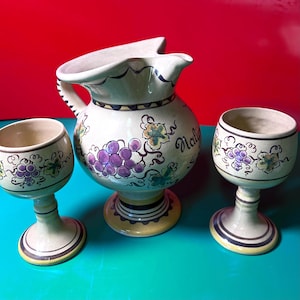 May include: A ceramic jug and two wine glasses featuring a grape design. The off-white jug has a spout and handle, with purple grapes and green leaves painted on the side. The glasses have a similar design and are on stems.