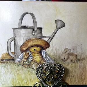 May include: A watercolour painting featuring a teddy bear in a straw hat, a watering can, and a rabbit. The bear holds a smaller teddy bear and flowers. A decorative heart-shaped object with rhinestones is in the foreground.