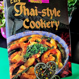 May include: A cookbook titled Easy Thai-style Cookery from the Australian Women's Weekly Home Library. The cover features a dark background with a statue and a bowl of shrimp curry with pineapple and asparagus, held by a heart-shaped metal stand.