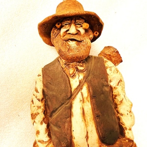 May include: A ceramic figurine of an elderly man with a long beard, wearing a hat and vest. The figurine is painted in shades of brown and tan, with details like the man's facial features and clothing. The figure has a jovial expression.