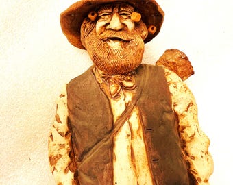 May include: A ceramic figurine of an elderly man with a long beard, wearing a hat and vest. The figurine is painted in shades of brown and tan, with details like the man's facial features and clothing. The figure has a jovial expression.