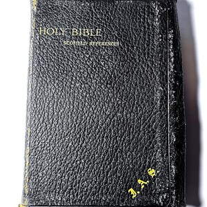 以下が含まれることがあります： 金色の文字が入った黒い革装丁の聖書。表紙には「HOLY BIBLE」と「SCOFIELD REFERENCES」の文字が見えます。端は摩耗しています。「J.A.S.」のイニシャルが右下に金色で印刷されています。本は使い込まれています。