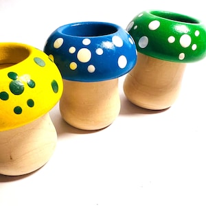 May include: Three wooden mushroom-shaped planters. The caps are painted yellow, blue, and green, each with white polka dots. The stems are a natural wood color. The planters are on a white background.