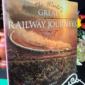 May include: A hardcover book titled The World's Great Railway Journeys by Tom Savio. The cover features a steam train travelling through a landscape of trees and hills, with a warm, golden sunset glow illuminating the scene.