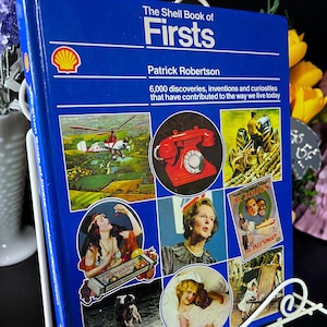 Może przedstawiać: Niebieska książka w twardej oprawie zatytułowana The Shell Book of Firsts autorstwa Patricka Robertsona, zawierająca logo Shell oraz różne historyczne obrazy, w tym helikopter, czerwony telefon z tarczą, traktor i portrety na okładce.