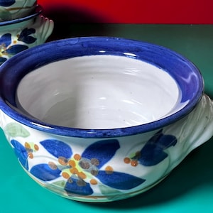May include: A white ceramic bowl with a blue rim and floral design. The bowl features a white interior and is decorated with blue flowers, orange accents, and green leaves. The bowl has a handle on the side.