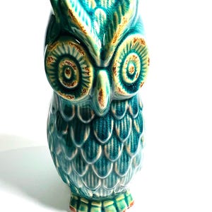May include: A teal ceramic owl ornament with brown and cream accents. The owl has large, detailed eyes and a textured surface, with a glossy finish. The figurine is standing upright on a white background.