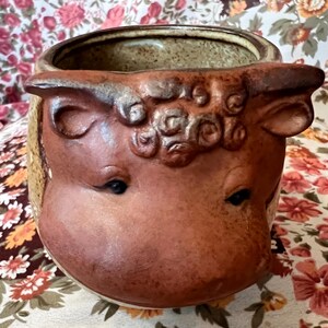Może przedstawiać: Ceramiczna donica z motywem twarzy krowy. Donica jest głównie brązowa z beżowymi i czarnymi akcentami. Twarz krowy jest wyrzeźbiona ze szczegółami, w tym oczami, uszami i teksturowanym czołem. Wnętrze jest w jasnobeżowym kolorze.