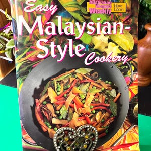 May include: A cookbook titled Easy Malaysian-Style Cookery from The Australian Women's Weekly Home Library. The cover features a black wok filled with stir-fried vegetables, including carrots, peas, and mushrooms, held by a heart-shaped metal stand.