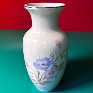 May include: A white ceramic vase with a silver rim and a floral design. The vase features a light blue flower with a yellow center, along with pink and purple flowers and green leaves. The vase has a classic, elegant shape.