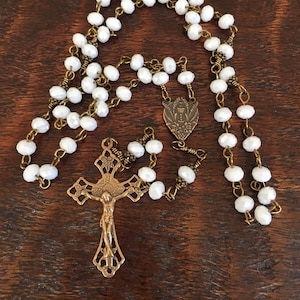 May include: A white crystal rosary with a brass chain and a crucifix. The crucifix features a detailed design and a figure of Jesus Christ. The centerpiece is a brass shield with a chalice and floral design.