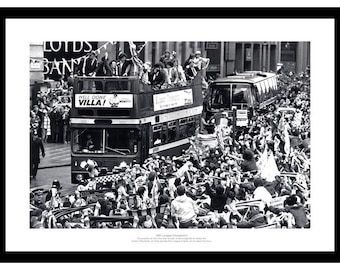 May include: Black and white photograph of a double-decker bus carrying a group of people, likely celebrating a victory. The bus is surrounded by a crowd of people waving flags and cheering. The words "WELL DONE VILLA!" are visible on the bus.