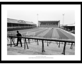 May include: Black and white photograph of a soccer stadium with a person walking on the field. The stadium has seating on both sides of the field. The field is green and has white lines. The sky is overcast.