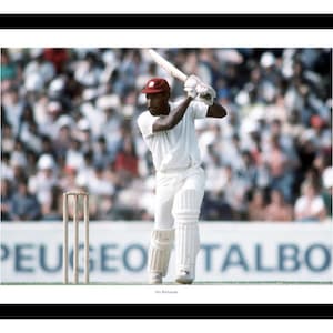 May include: A black and white photograph of a cricketer in a white uniform batting with a wooden bat. The player is wearing a red cap and is in a batting stance. The background is a blurred image of a crowd of people watching the game. The image has the text "Peugeot Talbot" in the background.