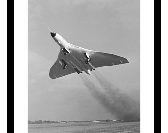 May include: A black and white photograph of an Avro Vulcan bomber taking off. The plane is in mid-air, with smoke trailing behind it. The plane is flying over a grassy field.