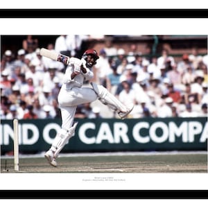 May include: A framed photograph depicting a cricket player mid-swing, wearing a white uniform and a red cap. The background shows a crowd and a green field. The image is framed in black, with the text 'Brian Lara' visible.