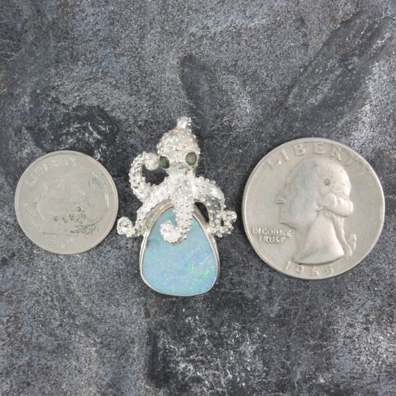 May include: Silver octopus pendant with a blue opal stone. The octopus has green eyes and is holding the opal stone. The pendant is about the size of a quarter.
