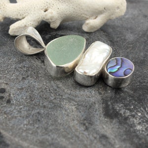 Puede incluir: Un colgante de plata con tres piedras: un vidrio de mar verde claro, una perla blanca y una concha de abul&oacute;n azul.