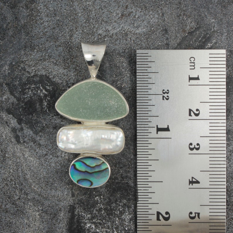 Puede incluir: Un colgante de plata con tres capas de diferentes piedras. La capa superior es una piedra verde claro, la capa intermedia es una piedra blanca y la capa inferior es una concha de abul&oacute;n azul y verde.