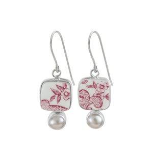 May include: A pair of silver drop earrings. Each earring features a square, white ceramic tile with a red floral pattern. A small, round pearl hangs below each tile. The earrings are set against a white background.