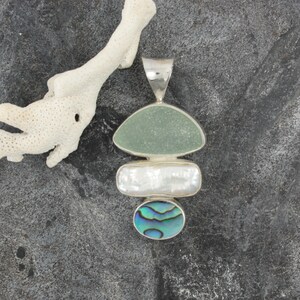 Puede incluir: Un colgante de plata con tres piedras superpuestas. La piedra superior es un vidrio de mar verde claro, la piedra del medio es una perla blanca y la piedra inferior es una concha de abul&oacute;n azul y verde.