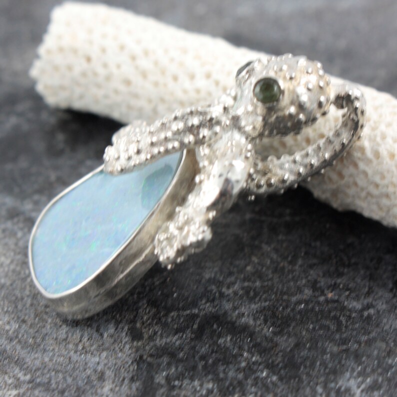 May include: A silver octopus pendant with a blue opal stone and green gemstone eyes. The octopus is detailed with textured tentacles and a textured body.