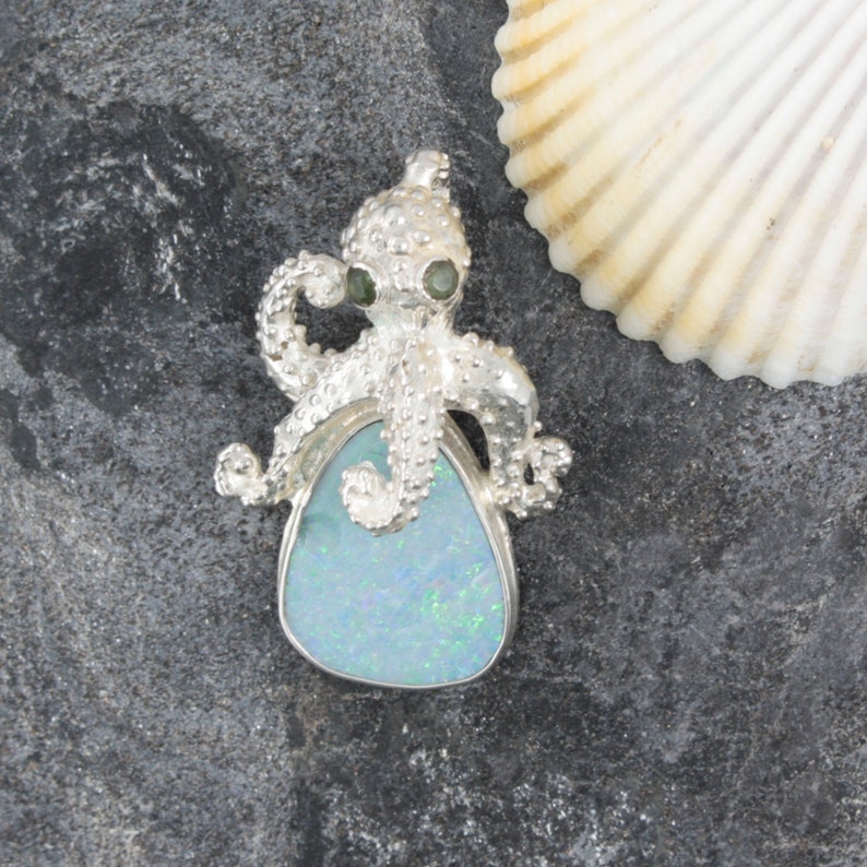 May include: A silver octopus pendant with green eyes and a blue opal stone. The pendant is set against a gray background with a seashell.