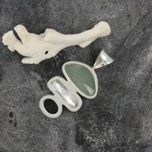 Puede incluir: Un colgante de plata con una piedra verde, una perla blanca y un anillo de plata. El colgante est&aacute; sobre un fondo gris oscuro.