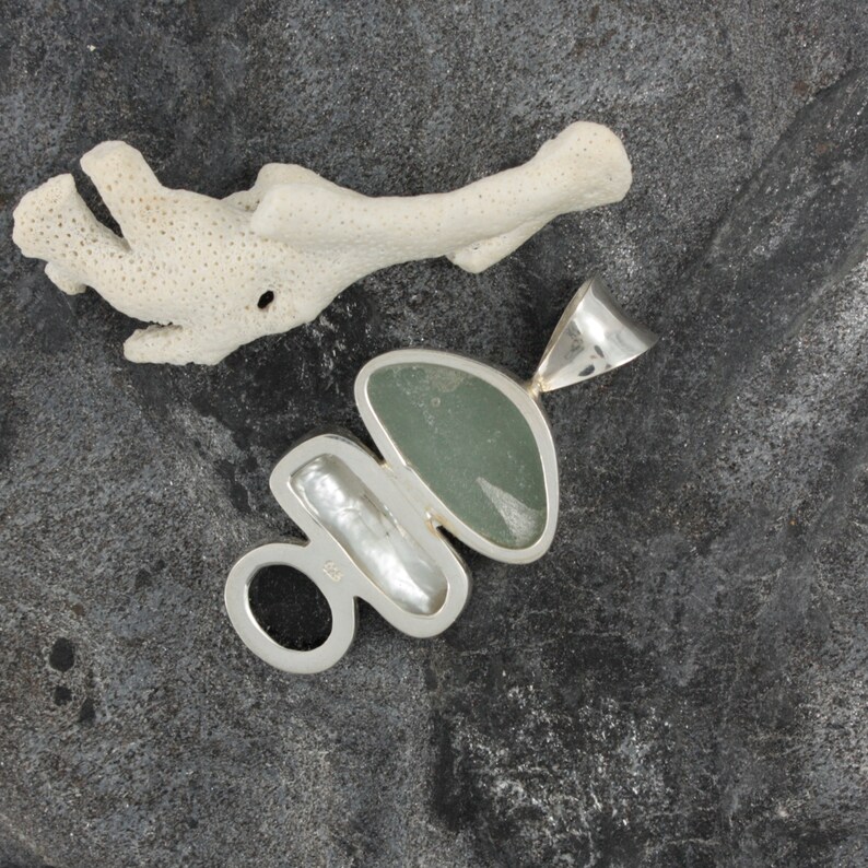 Puede incluir: Un colgante de plata con una piedra verde, una perla blanca y un anillo de plata. El colgante est&aacute; sobre un fondo gris oscuro.
