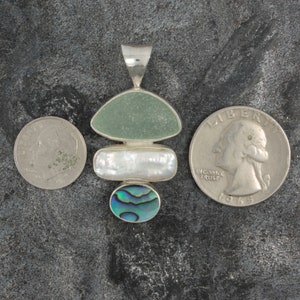Puede incluir: Un colgante de plata con tres piedras. La piedra superior es un vidrio de mar verde claro, la piedra del medio es una perla blanca y la piedra inferior es una concha de abul&oacute;n azul y verde.