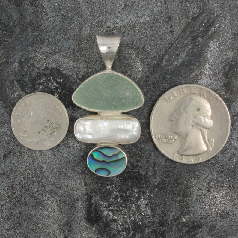 Puede incluir: Un colgante de plata con tres piedras. La piedra superior es un vidrio de mar verde claro, la piedra del medio es una perla blanca y la piedra inferior es una concha de abul&oacute;n azul y verde.