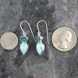May include: Sterling silver dangle earrings with a teardrop shaped blue lace agate gemstone and a smaller round blue topaz gemstone. The earrings are shown next to a US quarter for size comparison.