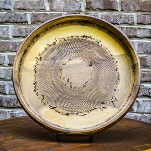Puede incluir: Una bandeja redonda de madera con interior marr&oacute;n claro y borde marr&oacute;n oscuro. El centro presenta un patr&oacute;n en espiral. La bandeja se exhibe sobre una mesa de madera oscura con un fondo de ladrillo.