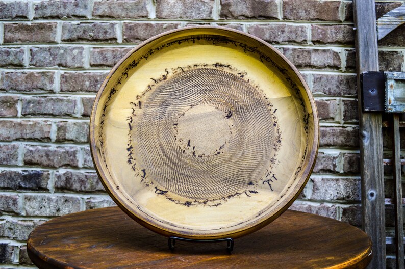 Puede incluir: Una bandeja redonda de madera con interior marr&oacute;n claro y borde marr&oacute;n oscuro. El centro presenta un patr&oacute;n en espiral. La bandeja se exhibe sobre una mesa de madera oscura con un fondo de ladrillo.