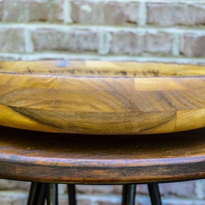 Puede incluir: Un cuenco de madera hecho a mano con un dise&ntilde;o ancho y poco profundo. El cuenco est&aacute; hecho de varias piezas de madera, mostrando patrones de vetas naturales y tonos c&aacute;lidos. Se encuentra sobre un taburete de madera oscura, con una pared de ladrillo al fondo.